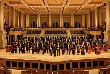 Os músicos da Orquestra estão posicionados na Estação das Artes da Sala São Paulo, um grande salão com colunas beges e chão com desenhos com de rosa. Os músicos vestem preto e estão segurando seus instrumentos.