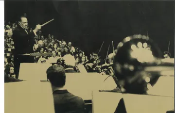 Eleazar de Carvalho em retrato preto e branco. Ele segura uma batuta com a mão direita e olha para a orquestra. Ao fundo, a plateia está lotada.