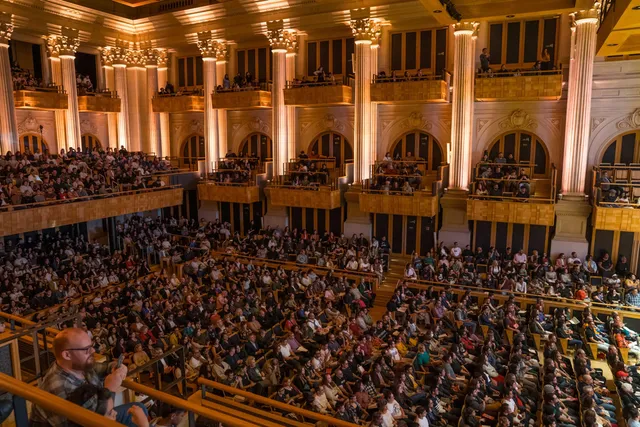 The concert hall is packed with people seated. The columns are illuminated with a yellow light.