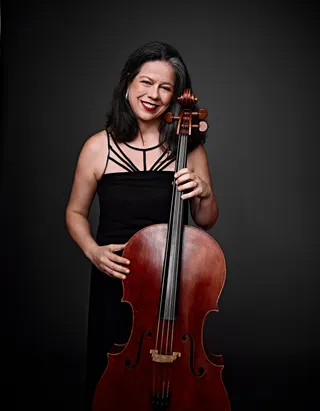 Adriana Holtz é uma mulher de cabelos pretos com uma mecha grisalha. Ela está usando um vestido preto e segurando um violoncelo.