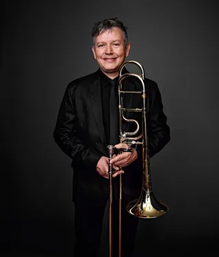 Alex Tartaglia is a white man with gray hair. He is wearing black clothing, smiling, and holding a trombone.