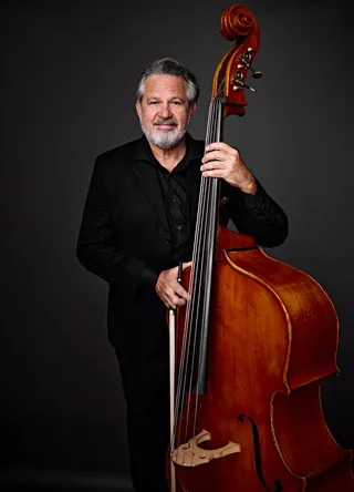 Alexandre Rosa é um homem branco, com cabelo e barba grisalhos. Ele está usando uma camisa preta e um paletó, e segura um contrabaixo.