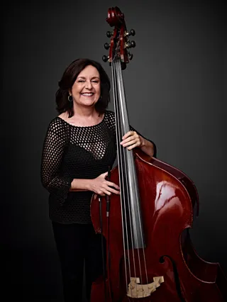 Ana Valéria Poles is a woman with shoulder-length brown hair. She is wearing a black crocheted dress and holding a double bass.