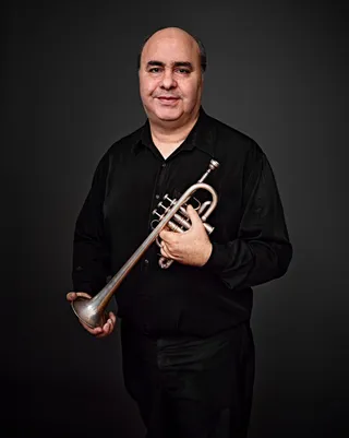Antonio Carlos Lopes Júnior is a bald white man. He is wearing a black shirt and holding a trumpet.