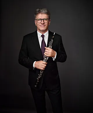 Arcadio Minczuk is a white man with gray hair and glasses. He is wearing a white shirt, a purple tie, a black jacket, and is holding an oboe.