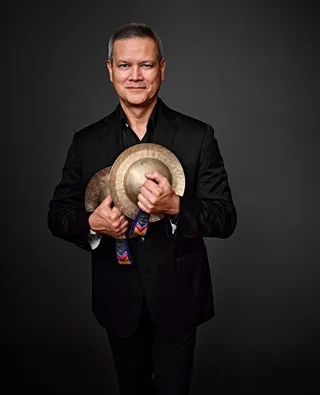 Armando Yamada is an Asian man with gray hair. He is holding two golden cymbals close to his body.