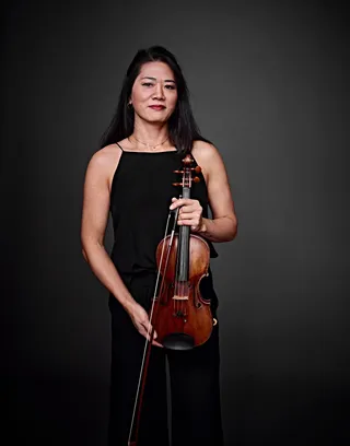 Camila Yasuda is an Asian woman with long black hair. She is wearing a black dress and holding a violin.