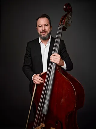 Claudio Torezan is a white man with brown hair and a gray beard. He is wearing a white shirt and a black suit jacket, and he is holding a double bass.