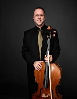Douglas Kier is a white man with short hair and a white beard. He is wearing a yellow tie, a black shirt, and a black suit jacket, and he is holding a cello.