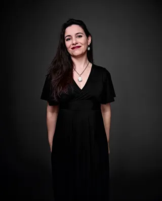 Fabiana Portas é uma mulher branca com cabelo preto e longo. Ela está usando um vestido preto e um conjunto combinando de colar e brincos com uma pedra verde-clara.