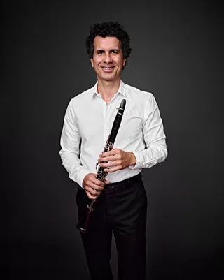 Giuliano Rosas is a white man with curly hair. He is smiling and holding a clarinet.