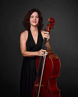 Heloisa Meirelles is a white woman with shoulder-length brown hair. She is wearing a low-cut black dress and holding a cello.