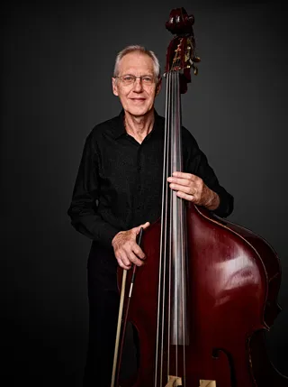 Max Ebert Filho is a white man with white hair, light-colored eyes, and glasses. He is wearing a black shirt and holding a double bass.