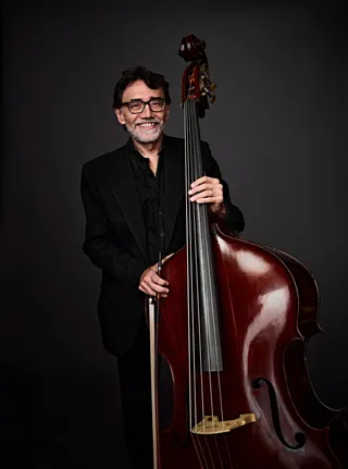 Ney Vasconcelos is a white man with black hair and a gray beard. He is wearing black glasses, a black suit jacket, and a black shirt, and he is holding a double bass.