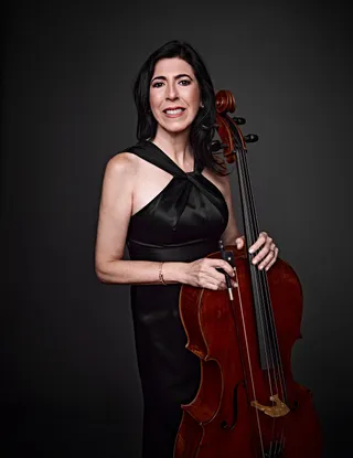 Regina Vasconcellos is a white woman with brown hair and brown eyes. She is wearing a strapless black dress and holding a cello.