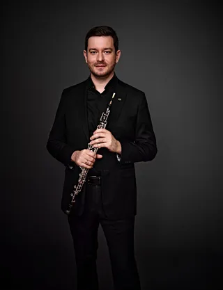 Ricardo Barbosa is a white man with brown hair and light-colored eyes. He is wearing a black shirt and blazer, and he is holding an oboe.