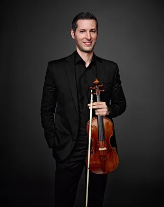 Rodolfo Lota is a white man with black hair and beard. He is wearing a black shirt and suit jacket, and he is holding a violin.