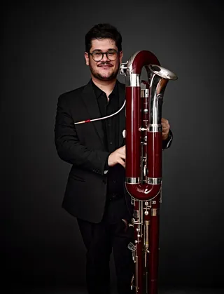 Romeu Rabelo é um homem branco, de cabelo preto, barba e bigode pretos, usando óculos redondos. Ele está vestido com uma camisa preta e segurando um fagote.