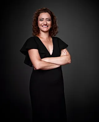 Silvana Romani é uma mulher branca, com cabelo loiro e cacheado. Ela está usando um vestido preto e sorrindo.