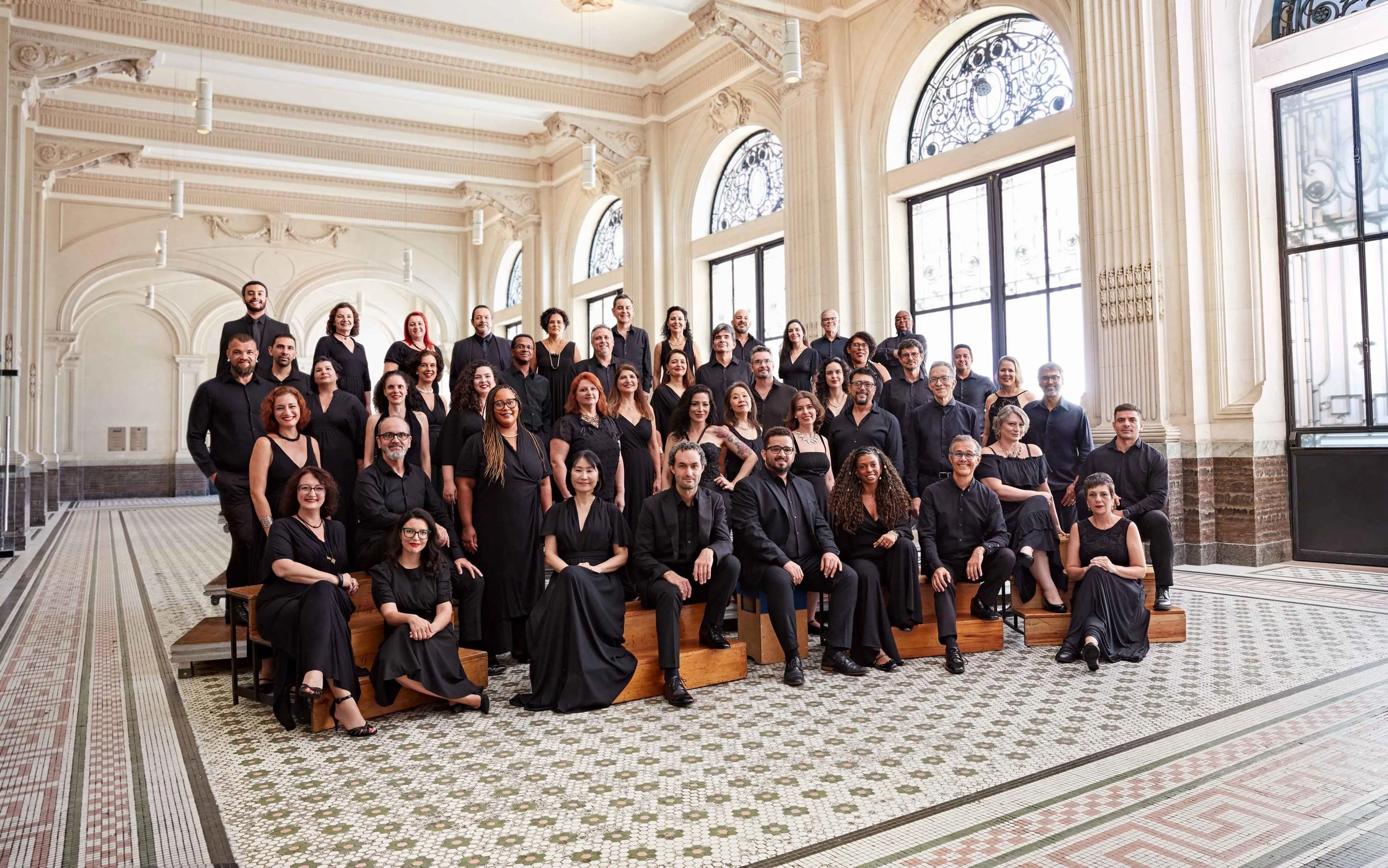 Os músicos do Coro estão posicionados no Hall Principal da Sala São Paulo, todos vestem preto e sorriem.