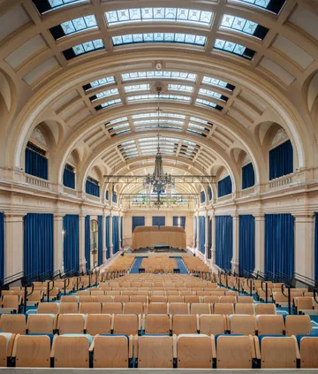 Vista ampla da Estação Motiva Cultural, com piso em tom rosado e paredes beges. Ao fundo, três grandes portões metálicos e um vitral colorido. Nas laterais, grandes arcos com estruturas de vidro e metal. O teto é oval e contra com um lustre ao centro.