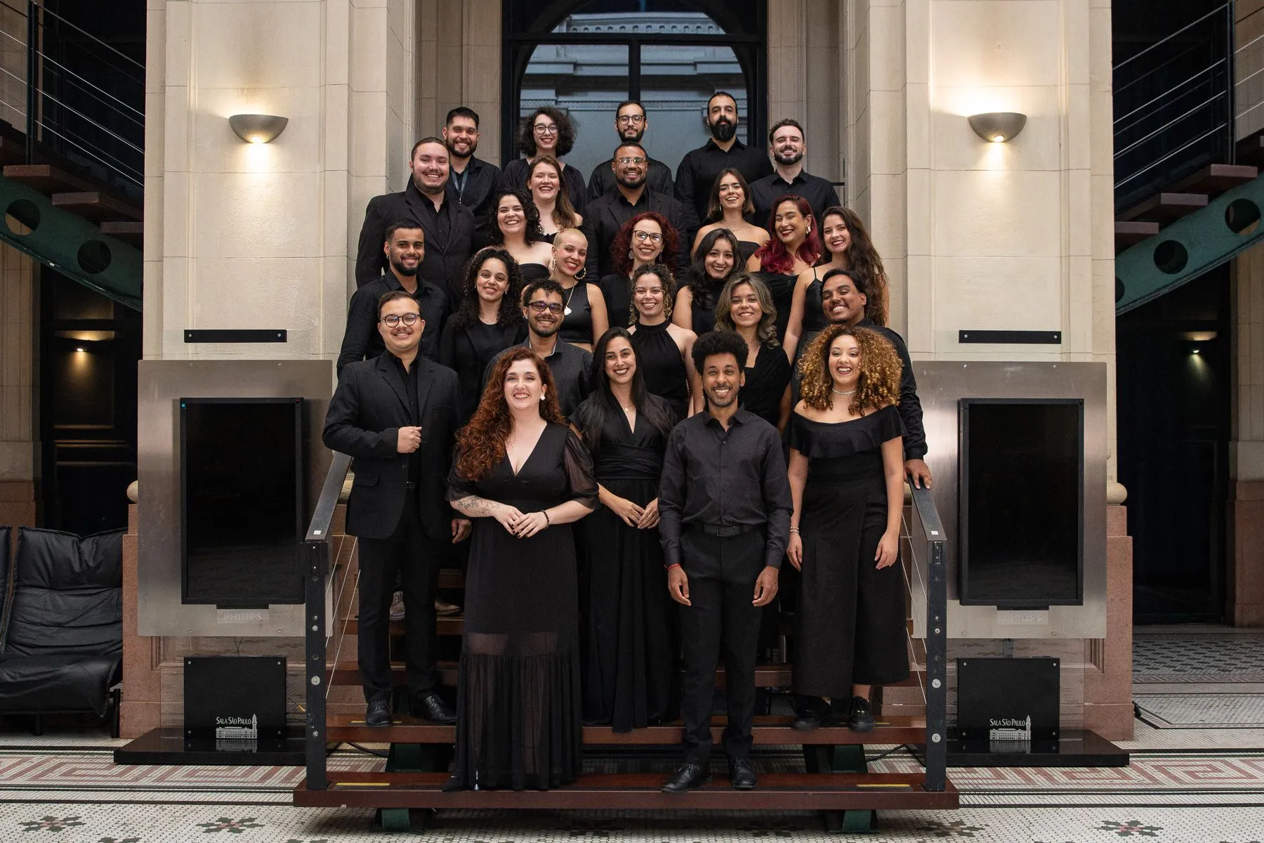 Os coristas da Academia de Música estão posicionados em uma escada no Salão São Paulo. Todos estão vestindo roupas pretas e sorrindo.