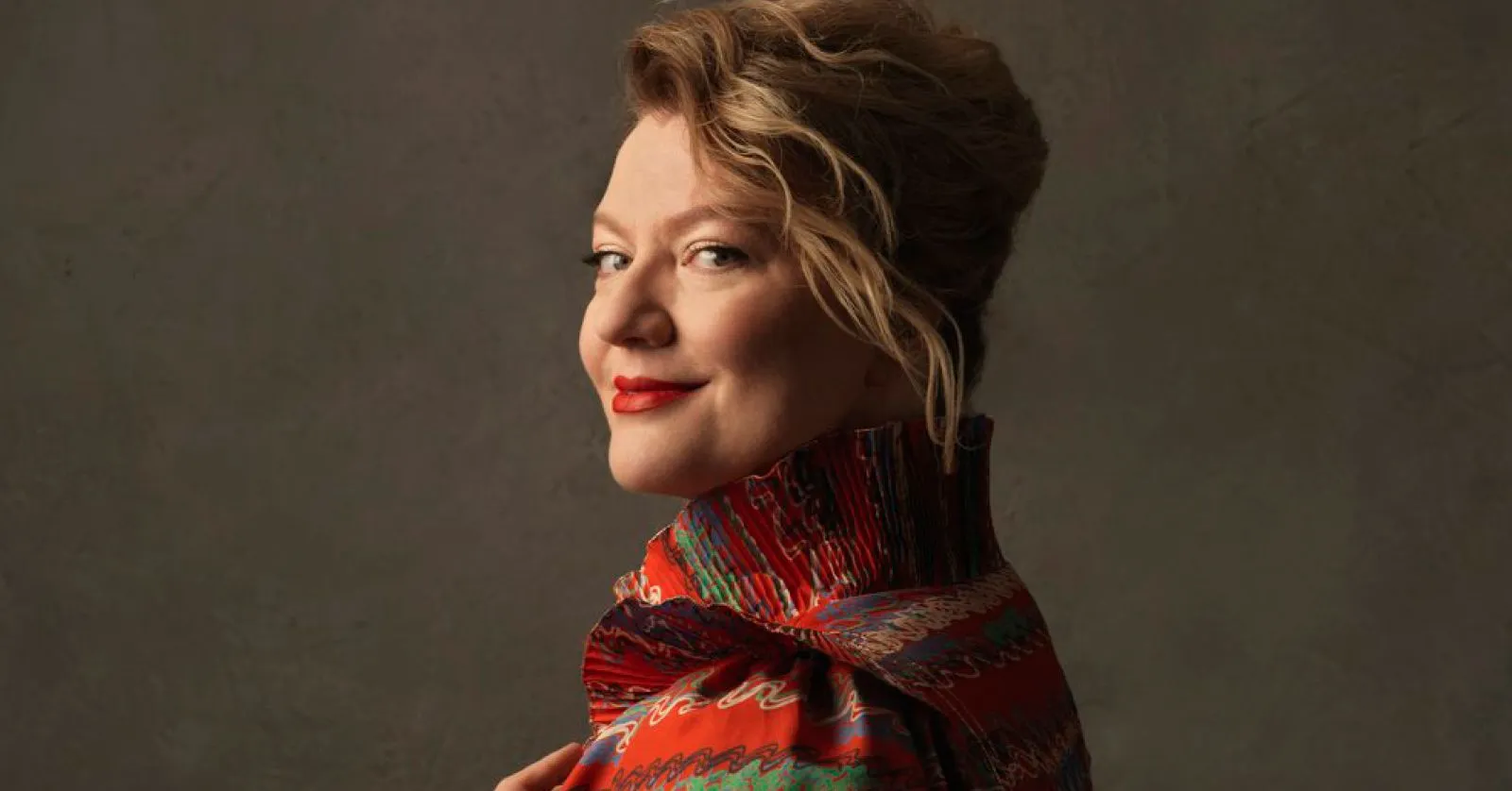 Aile é uma mulher loira, branca, que usa uma roupa estampada vermelha e um batom também vermelho.