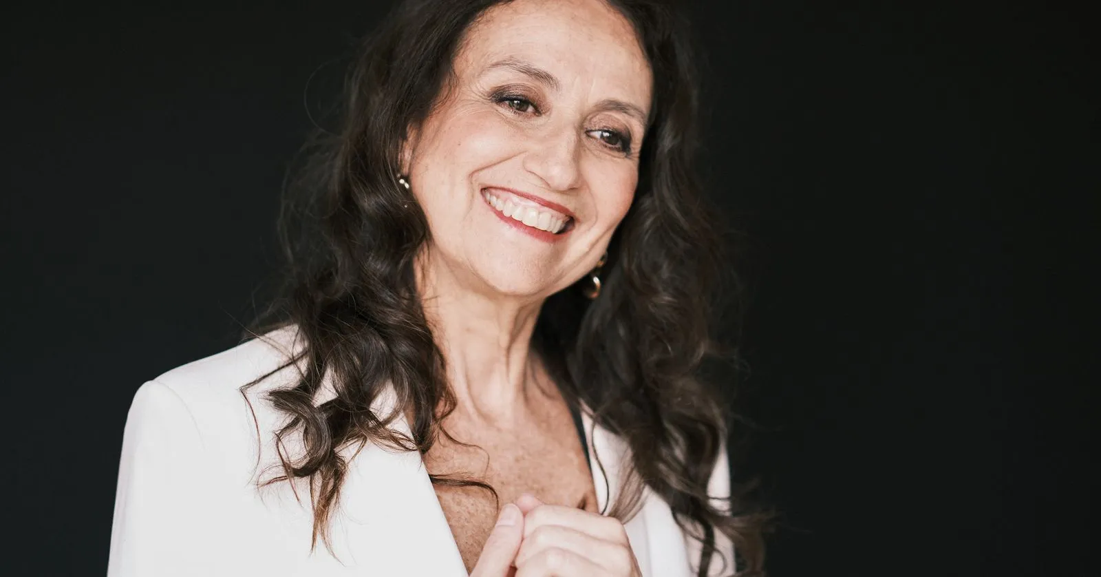 Sonia Rubinsky é uma mulher branca, de cabelos castanhos e longos. Ela veste branco e sorri com a mão no queixo.