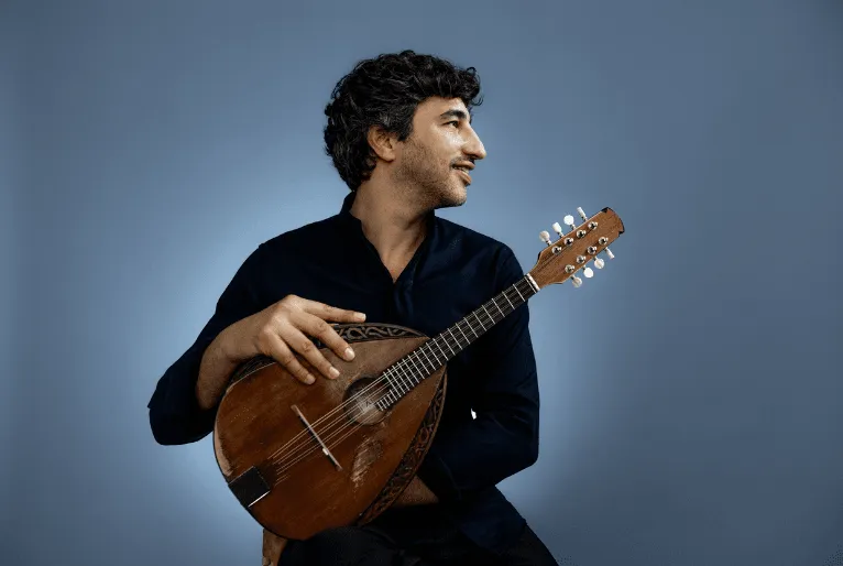 Foto de Avi Avital, homem branco e cabelos castanhos escuros. Ele veste camisa preta, segura um bandolim, instrumento de cordas, com uma das mãos a frente do corpo e olha para o lado.