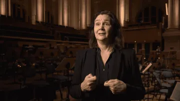 Foto da contralto Anna Larsson, mulher branca de cabelos pretos e compridos. Ela usa camisa social preta de mangas longas e está dentro da Sala São Paulo.