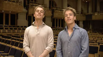 Foto de  Arthur Jussen e Lucas Jussen. Arthur está na esquerda, é um homem branco, loiro e veste uma camiseta bege com golas abertas. Lucas está na direita, é um homem branco, de cabelos loiros e veste uma camisa azul de botões.