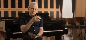 Foto de Sir Richard Armstrong, homem branco e de cabelos brancos. Ele está sentado, veste uma camisa de mangas curtas preta e usa óculos retangulares. Está sentado a frente de um piano.