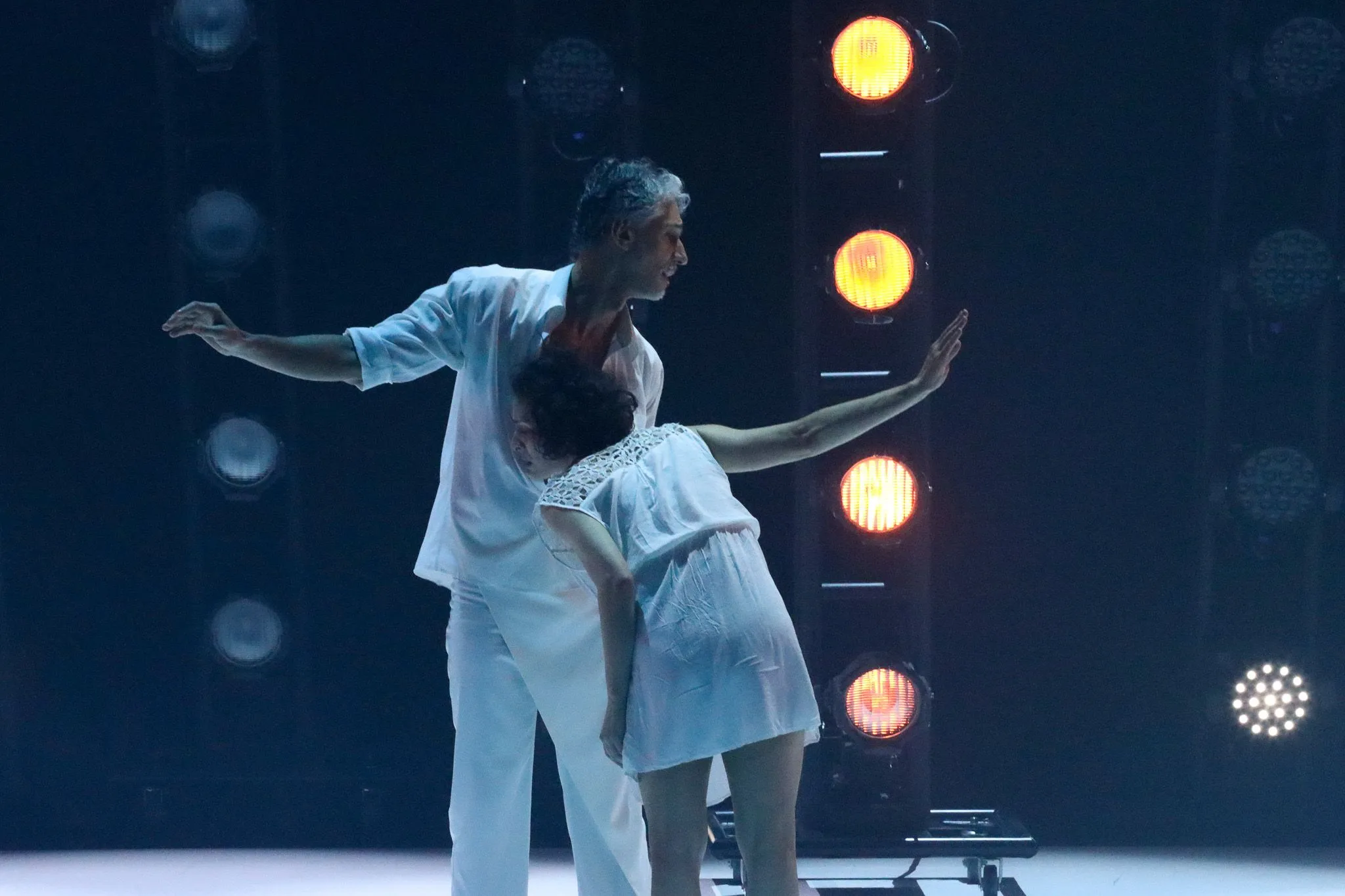 Dois dançarinos no palco. Eles usam roupa branca e no fundo temos luzes acesas.