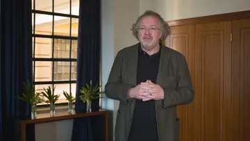 Stéphane Denève está em pé em um ambiente interno e as mãos entrelaçadas. Ela veste blazer cinza, camisa preta e calça jeans.