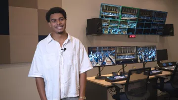 Foto de Samuel Alves, homem preto, veste camisa branca de mangas curtas. Ele está de pé dentro de um estúdio de gravação, ao fundo é possível ver algumas telas com imagens da sala de cocnertos.