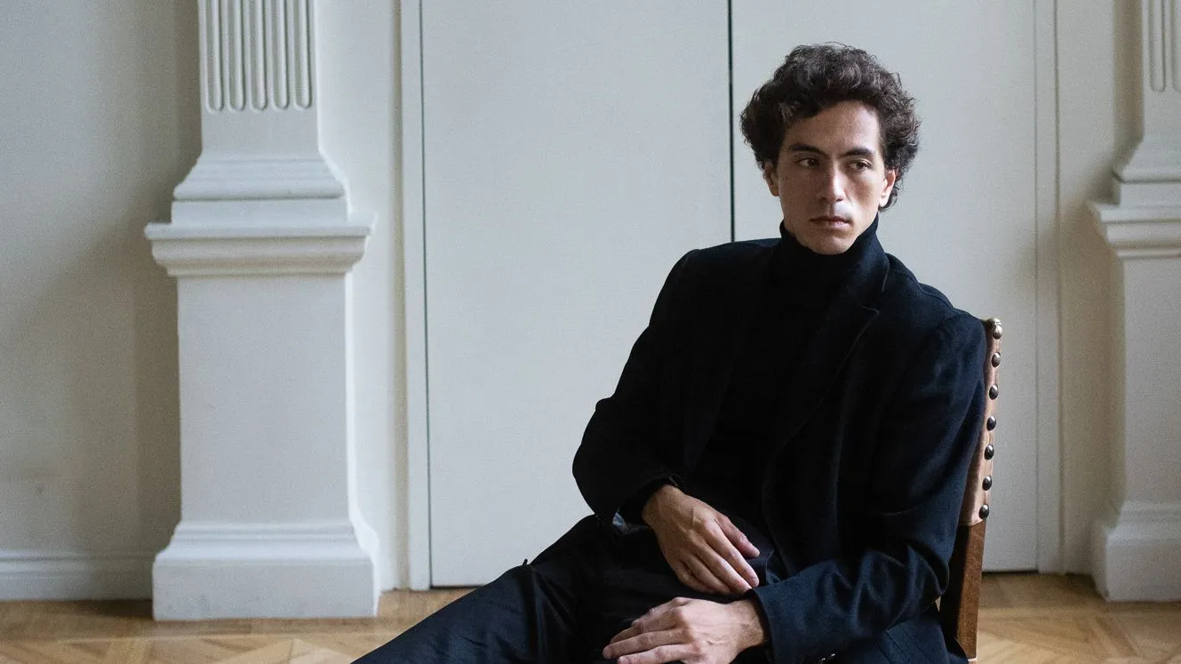 Konstantin Emelyanov, um homem branco vestindo preto, sentado em uma cadeira de madeira em uma sala branca.