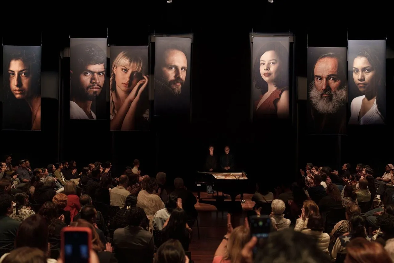 Sala de concertos ampla e vazia, com fileiras de cadeiras de madeira voltadas para um palco central. O espaço tem arquitetura clássica, pé-direito alto, grandes cortinas azuis nas laterais e um lustre no teto. Ambiente interno bem iluminado.