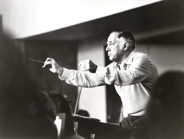 Eleazar de Carvalho é um homem branco, calvo. Ele está regendo a orquestra.