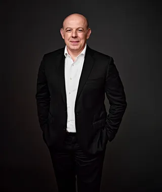Nikolay Genov é um homem branco, calvo. Ele sorri, veste uma camisa branca e paletó preto, e segura uma trompa.