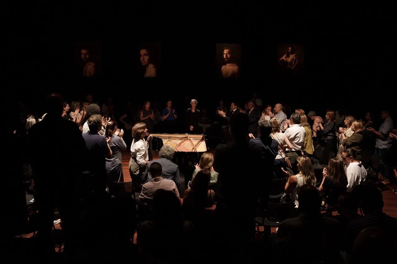 Sala de concertos ampla e vazia, com fileiras de cadeiras de madeira voltadas para um palco central. O espaço tem arquitetura clássica, pé-direito alto, grandes cortinas azuis nas laterais e um lustre no teto. Ambiente interno bem iluminado.