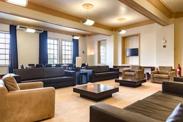 A Sala Camargo Guarnieri tem diversos sofás e poltronas delimitando o espaço interno, uma telwevisão na parede do fundo e janelas retangulares com cortinas azul marinho. As paredes são pintadas em tons de bege.