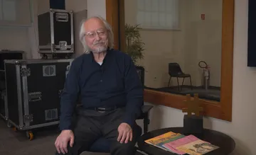 Masaaki Suzuki aparece sentado, com cabelos brancos longos e lisos, barba e bigode curtos também brancos. Usa óculos de armação fina e está vestido de camisa social escura com calça preta, transmitindo um ar sereno e elegante.