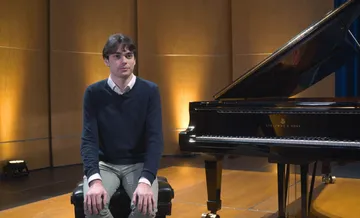 Tom Borrow aparece sentado em frente a um piano de cauda, em um palco iluminado. Ele tem cabelo castanho curto, pele clara e veste camisa clara com suéter azul-escuro e calça clara. Sua postura é formal e atenta, transmitindo serenidade e concentração.