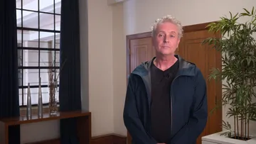 Foto de Alexander Liebreich, homem branco e de cabelos curtos e brancos. Ele veste camiseta preta e um casaco aberto azul escuro. No fundo, é possível ver um grande porta, ao lado direito, uma planta verde e ao lado esquerdo uma janela.