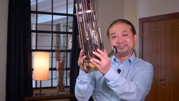 Foto de Wu Wei. Homem amarelo, de cabelos compridos e castanhos. Usa camisa azul claro. Ele segura um sheng, instrumento de sopro.