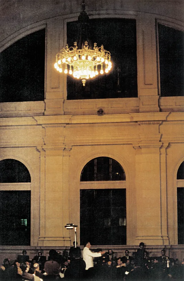 A foto mostra a Osesp tocando em um salão com um grande lustre, janelas e cortinas azuis.