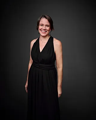 Ana Ganzert é uma mulher branca, de cabelo curto. Ela está usando óculos, um vestido preto e está sorrindo.