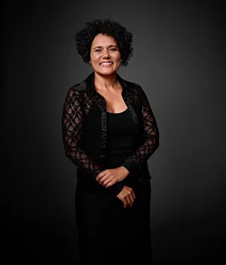 Anna Carolina é uma mulher de pele morena, com cabelo curto e cacheado. Ela está usando uma blusa de renda preta sobre uma regata preta. Ela usa batom nude e está sorrindo na foto.