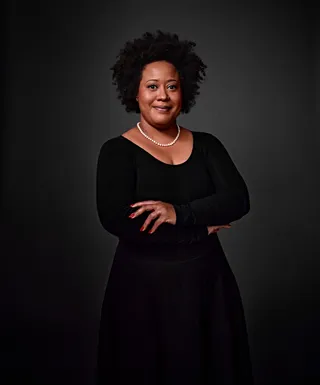 Erika Muniz is a Black woman. She is wearing a black dress, a pearl necklace, and has a piercing on the right side of her nose.