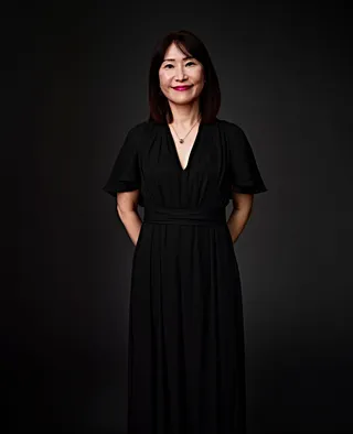 Ji Sook Chang é uma mulher coreana com cabelo na altura dos ombros e franja. Ela está usando batom rosa, um vestido preto e um colar de ouro com um pingente.
